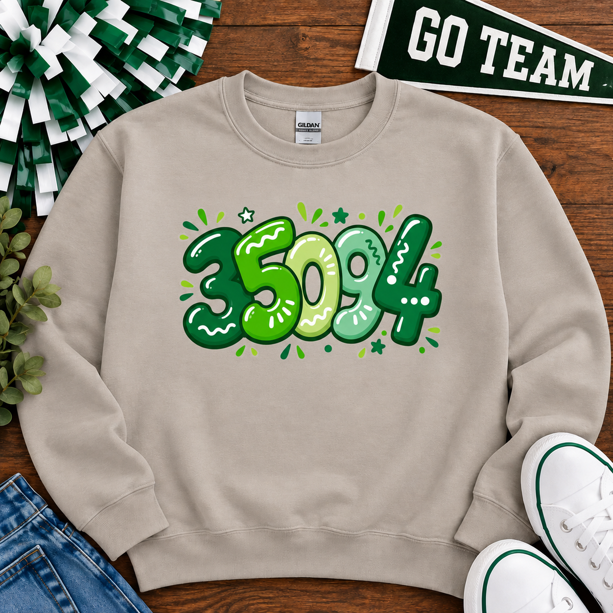 A beige crewneck sweatshirt featuring a green and light green '35094' graphic with shamrock accents, styled in a flat lay on a wooden surface with a pom-pom, jeans, white sneakers, and a 'GO TEAM' banner.