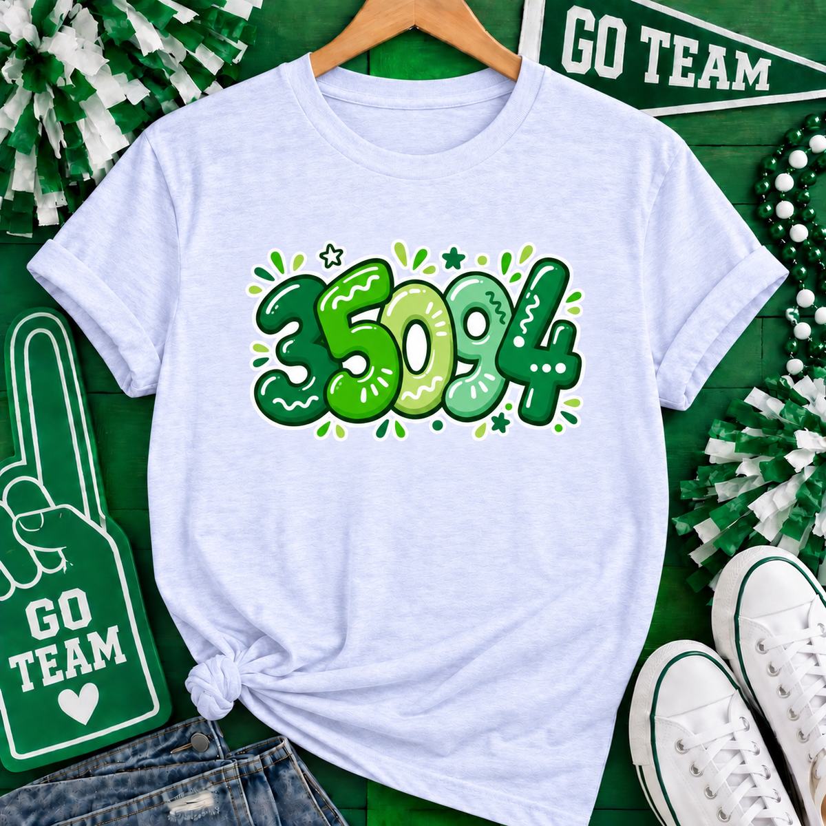 A light heather blue graphic t-shirt on a wooden hanger, styled in a flat lay with St. Patrick's Day themed props including green and white pom-poms, a foam finger sign, a beaded garland, white sneakers, and denim jeans.