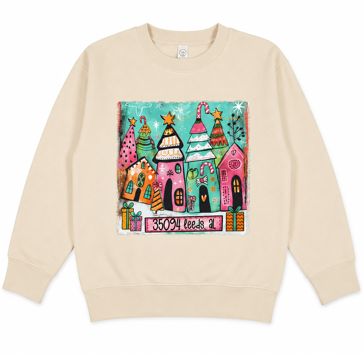 Cream crewneck sweatshirt featuring a colorful whimsical Christmas village design with festive houses, decorated trees, presents, and the text "35094 Leeds, AL" centered on the front.