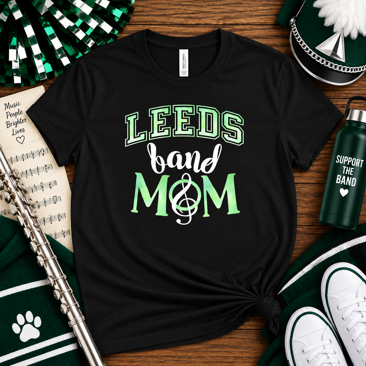 A black graphic t-shirt with the text 'LEEDS band MOM' in green and white, styled in a flat-lay arrangement with a silver flute, a green and white striped knit, a green water bottle, and a green pom-pom on a wooden background.