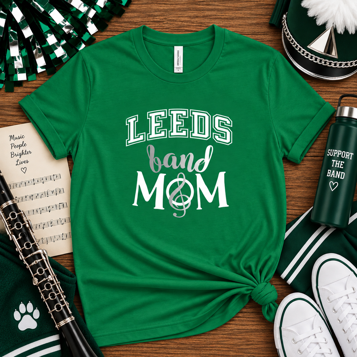 A green graphic t-shirt with 'LEEDS band MOM' printed in white, styled in a flat lay with school band props including a clarinet, shoes, a water bottle, and a pom-pom on a wooden background.