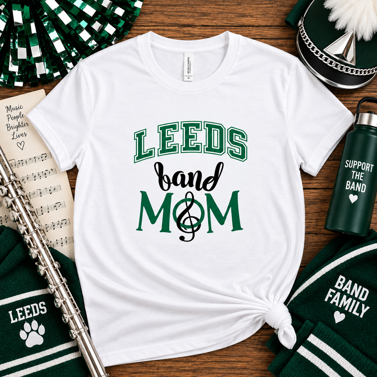A white graphic t-shirt with 'LEEDS band MOM' printed in green and black, styled in a flat lay with school band props including a flute, pom-poms, a water bottle, and a beanie on a wooden background.