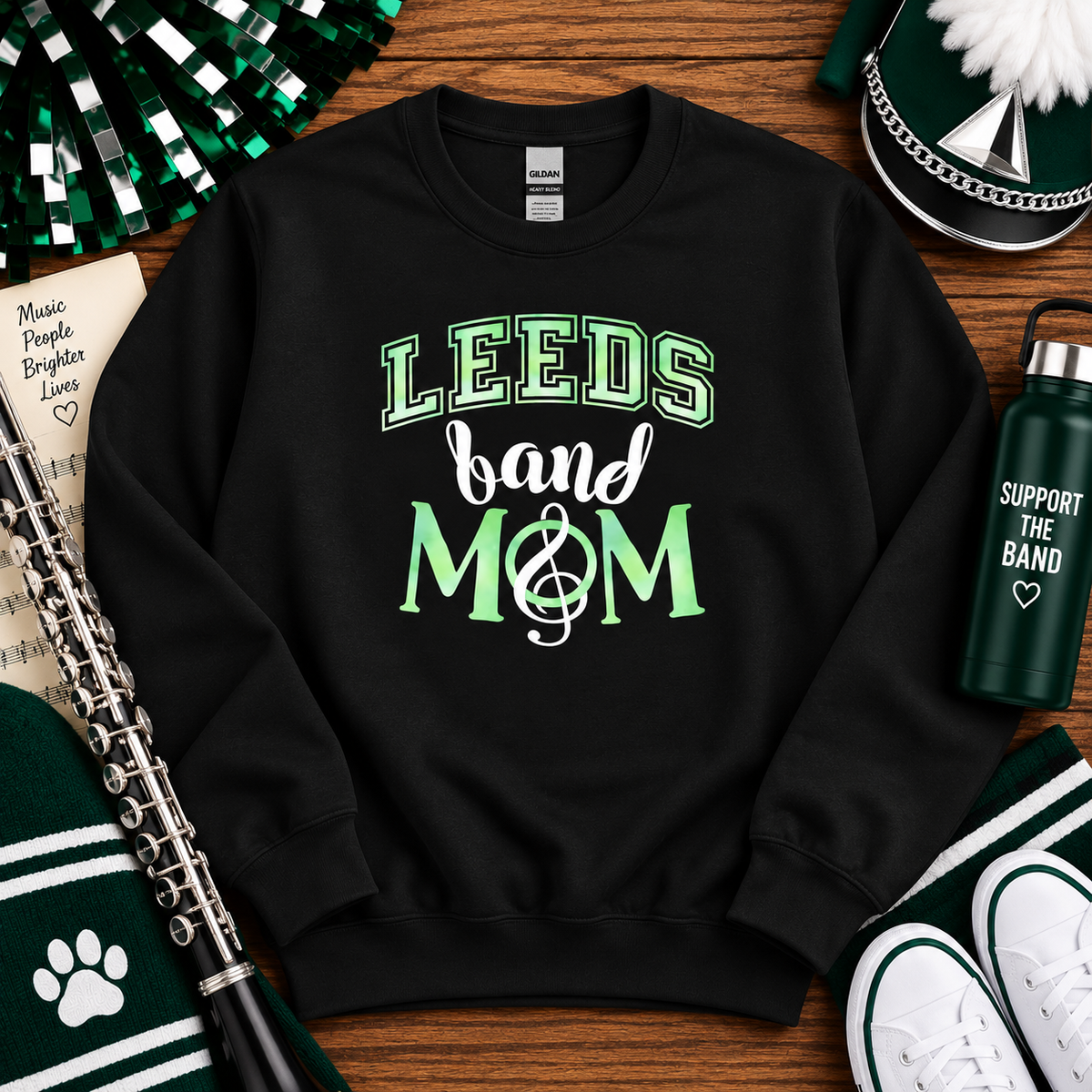A black crewneck sweatshirt with 'LEEDS band MOM' printed on the front in green and white, styled in a flat lay with St. Patrick's Day themed props including a green water bottle, pom-poms, a clarinet, and white sneakers on a wooden background.