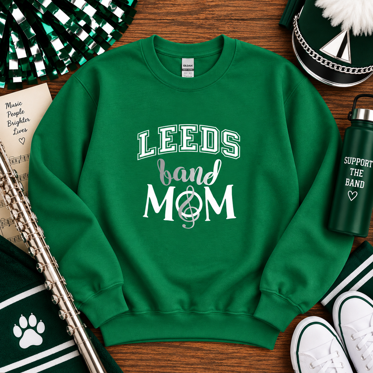 A green crewneck sweatshirt with 'LEEDS band MOM' printed in white on the front, styled in a flat lay on a wooden table with band-themed accessories including a flute, water bottle, and drum head.