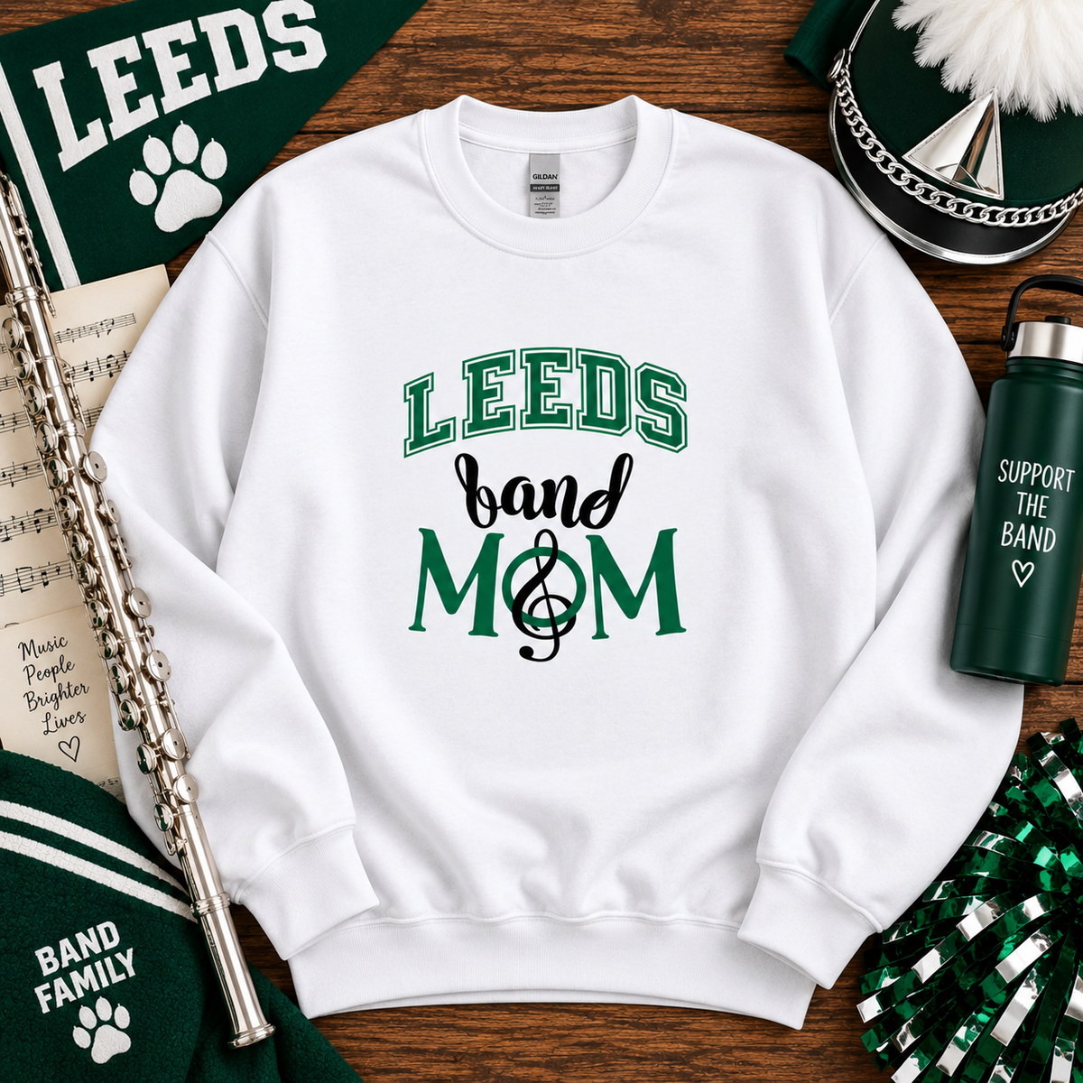 A white crewneck sweatshirt with a green and black graphic reading 'LEEDS band & MOM' with a treble clef, styled in a flat lay on a wooden surface with band-themed props including a flute, water bottle, pom-poms, and a hat.