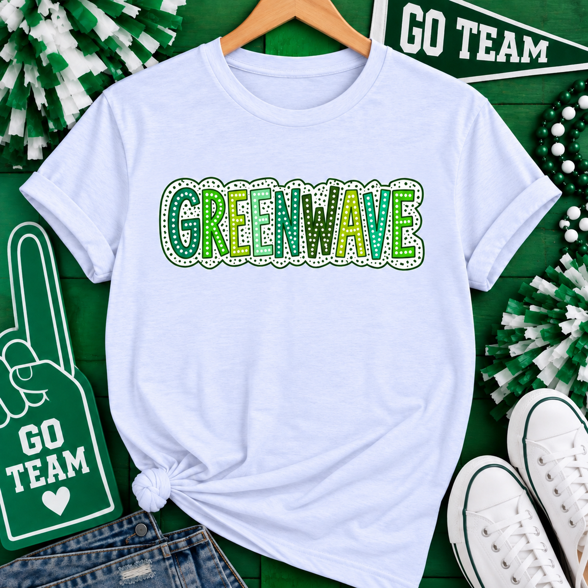 A white short-sleeve T-shirt on a wooden hanger, featuring a 'GREENWAVE' graphic across the chest in green and yellow with a dotted border. The shirt is styled in a flat lay on a green background with 'GO TEAM' signs, pom-poms, jeans, and white sneakers.