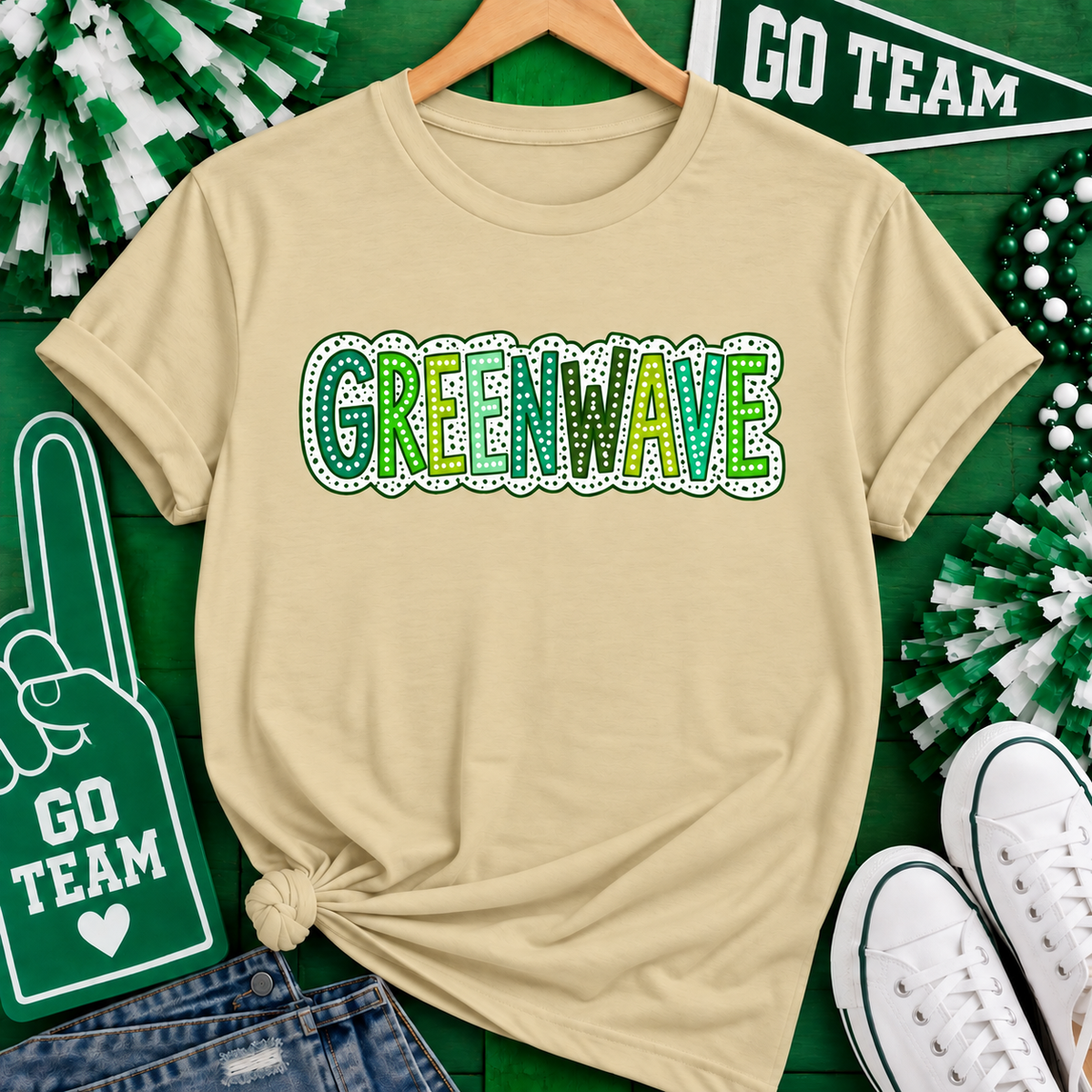 A beige short-sleeve T-shirt on a wooden hanger, featuring the word 'GREENWAVE' printed across the chest in green and white. The shirt is styled in a flat-lay arrangement with St.