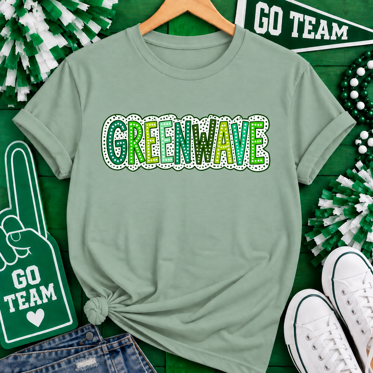 A sage green graphic t-shirt on a wooden hanger, featuring the word 'GREENWAVE' in green and yellow with a white dotted outline. The shirt is styled in a flat lay with St.