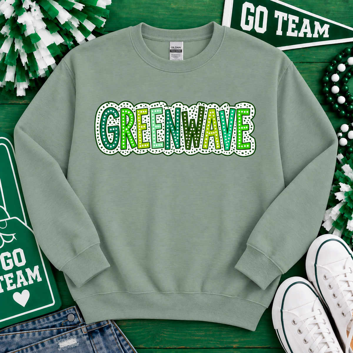 A sage green crewneck sweatshirt featuring the word 'GREENWAVE' in a glittery green and yellow font with a white dotted outline. The sweatshirt is styled in a flat-lay arrangement on a green wooden background with St.