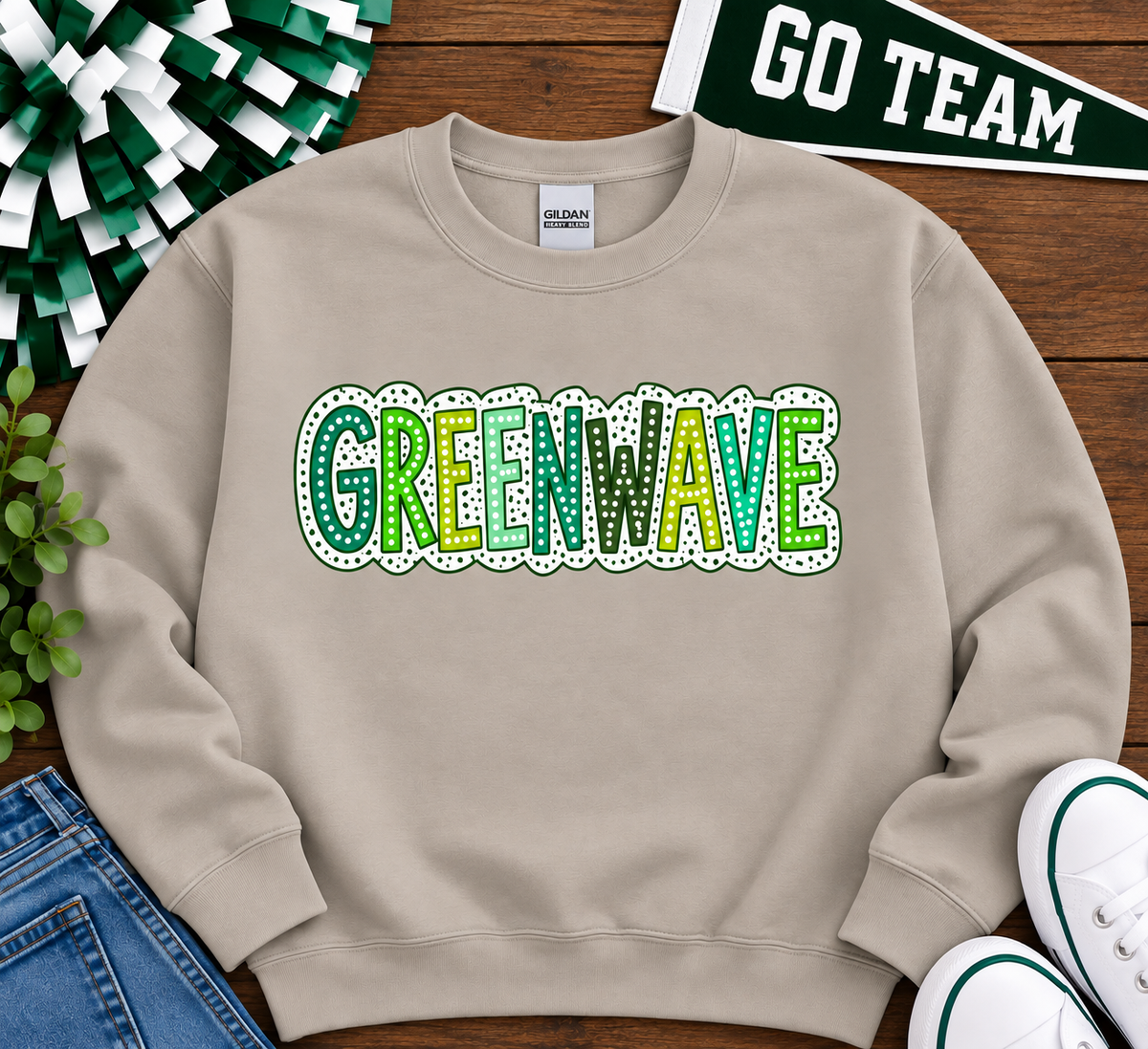 A beige crewneck sweatshirt with 'GREENWAVE' printed across the chest in green and yellow patterned letters with a white dotted border. The sweatshirt is displayed in a flat-lay arrangement on a wooden surface alongside a green and white pom-pom, a 'GO TEAM' banner, blue jeans, and white sneakers.