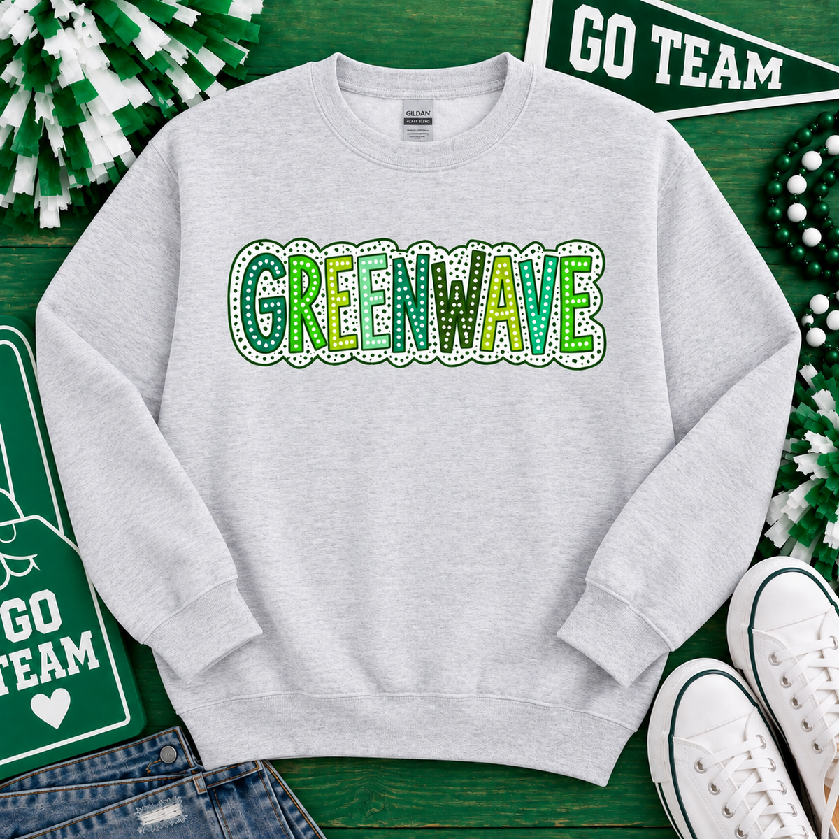 An ash sweatshirt with 'GREENWAVE' printed across the chest in green and yellow lettering with a white dotted border. The sweatshirt is styled in a flat-lay arrangement on a green wooden background with St.