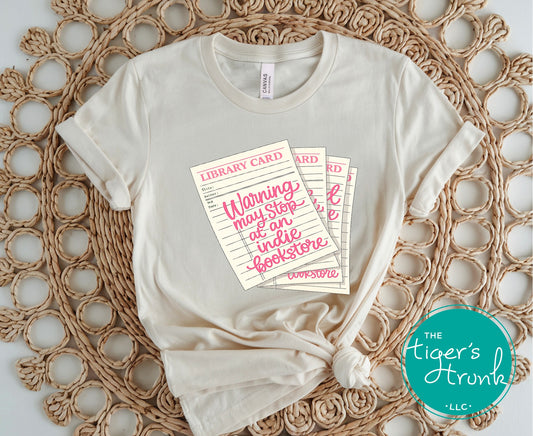 Library card shirt with warning may stop at an indie bookstore design on cream, navy, mauve, sage, sand, fatigue green, and maroon shirts