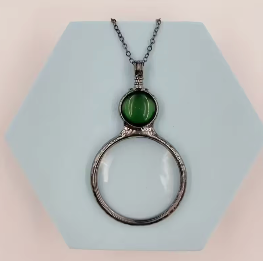 Magnifying glass pendant necklace with green accent stone and silver-tone chain displayed on a geometric background