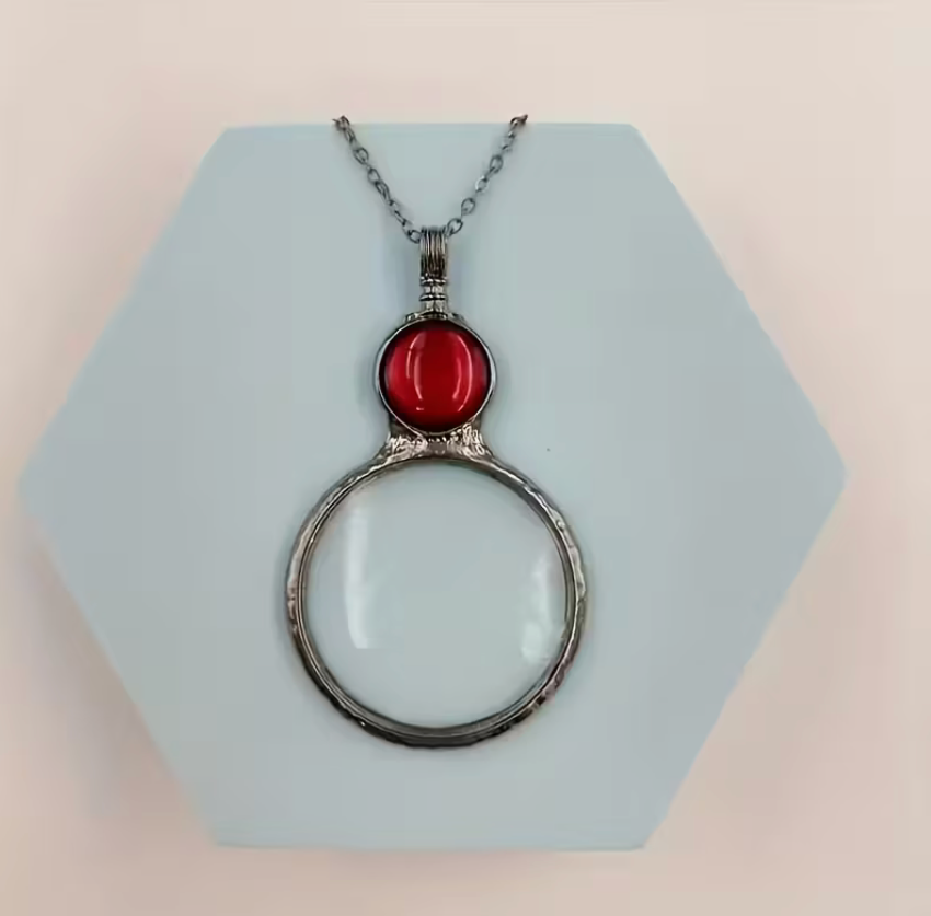 Magnifying glass pendant necklace with red accent stone and silver-tone chain displayed on a geometric background