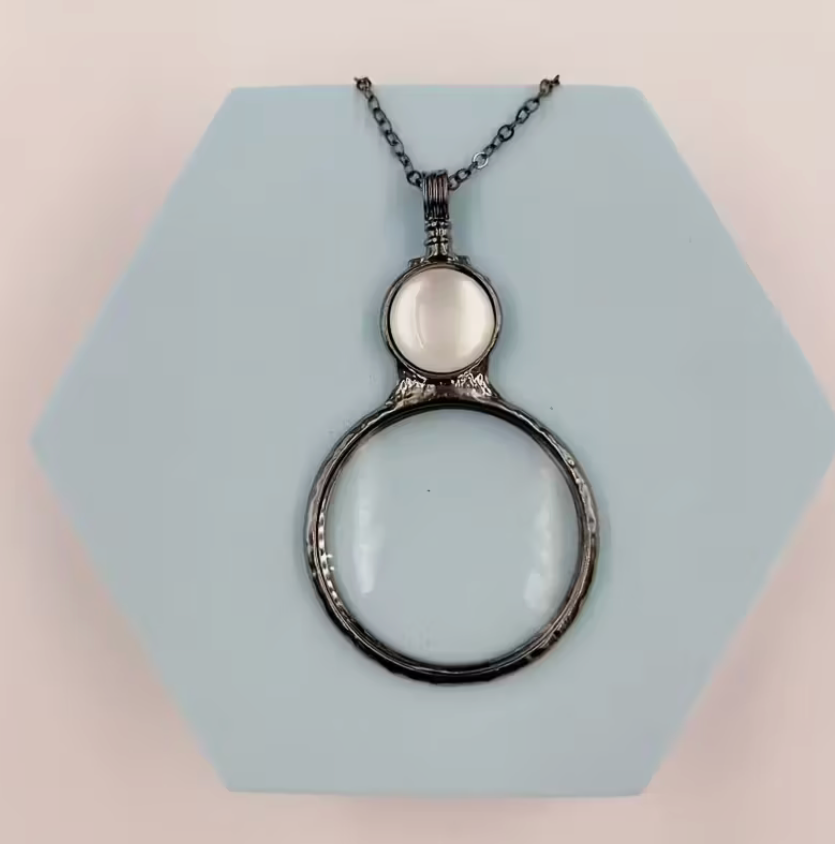 Magnifying glass pendant necklace with white accent stone and silver-tone chain displayed on a geometric background.