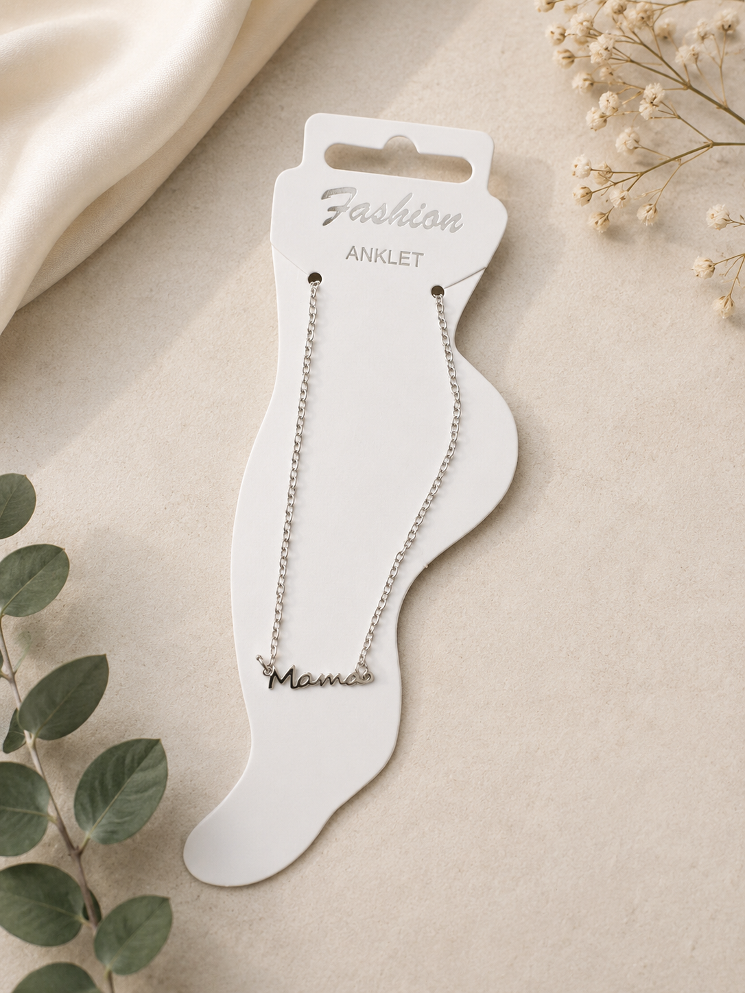 Silver mama anklet displayed on a foot-shaped jewelry card against an elegant neutral flat lay background with soft fabric, greenery, and delicate floral accents
