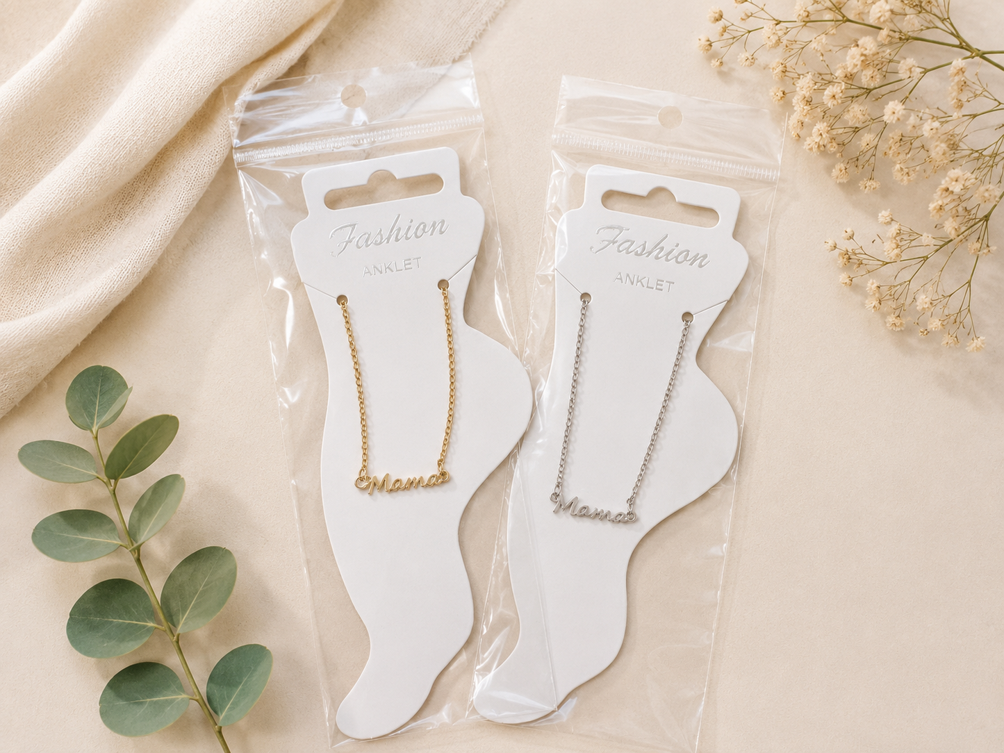 ackaged gold and silver mama anklets displayed on foot-shaped jewelry cards against an elegant neutral flat lay background with soft fabric, greenery, and delicate floral accents