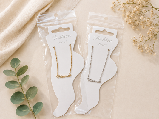 ackaged gold and silver mama anklets displayed on foot-shaped jewelry cards against an elegant neutral flat lay background with soft fabric, greenery, and delicate floral accents