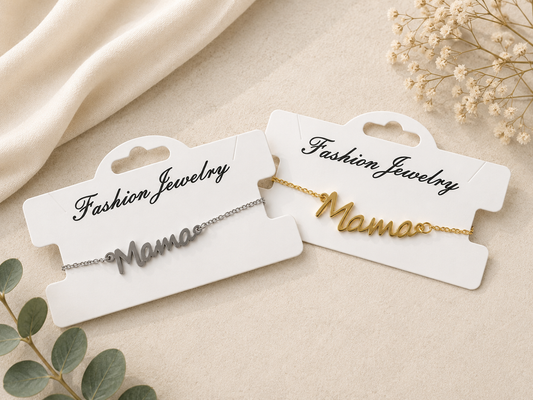 Gold and silver mama script bracelets displayed on white jewelry cards against an elegant neutral flat lay background with soft fabric, greenery, and delicate floral accents