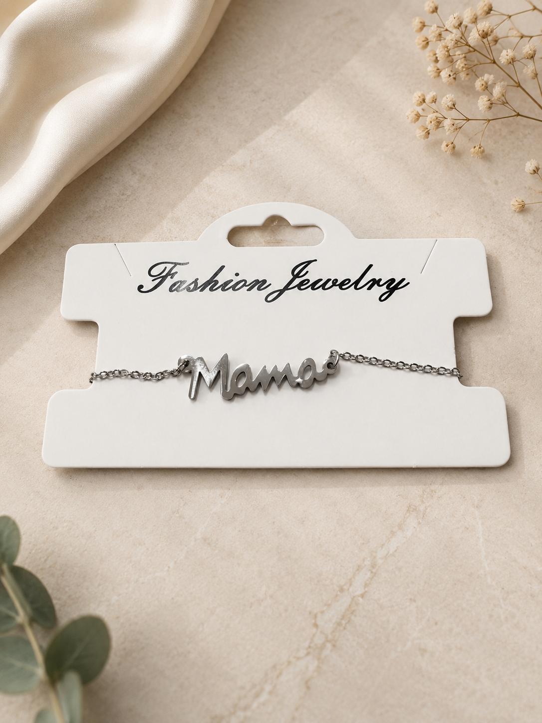 Silver mama script bracelet displayed on a white jewelry card against an elegant neutral flat lay background with soft fabric, greenery, and delicate floral accents
