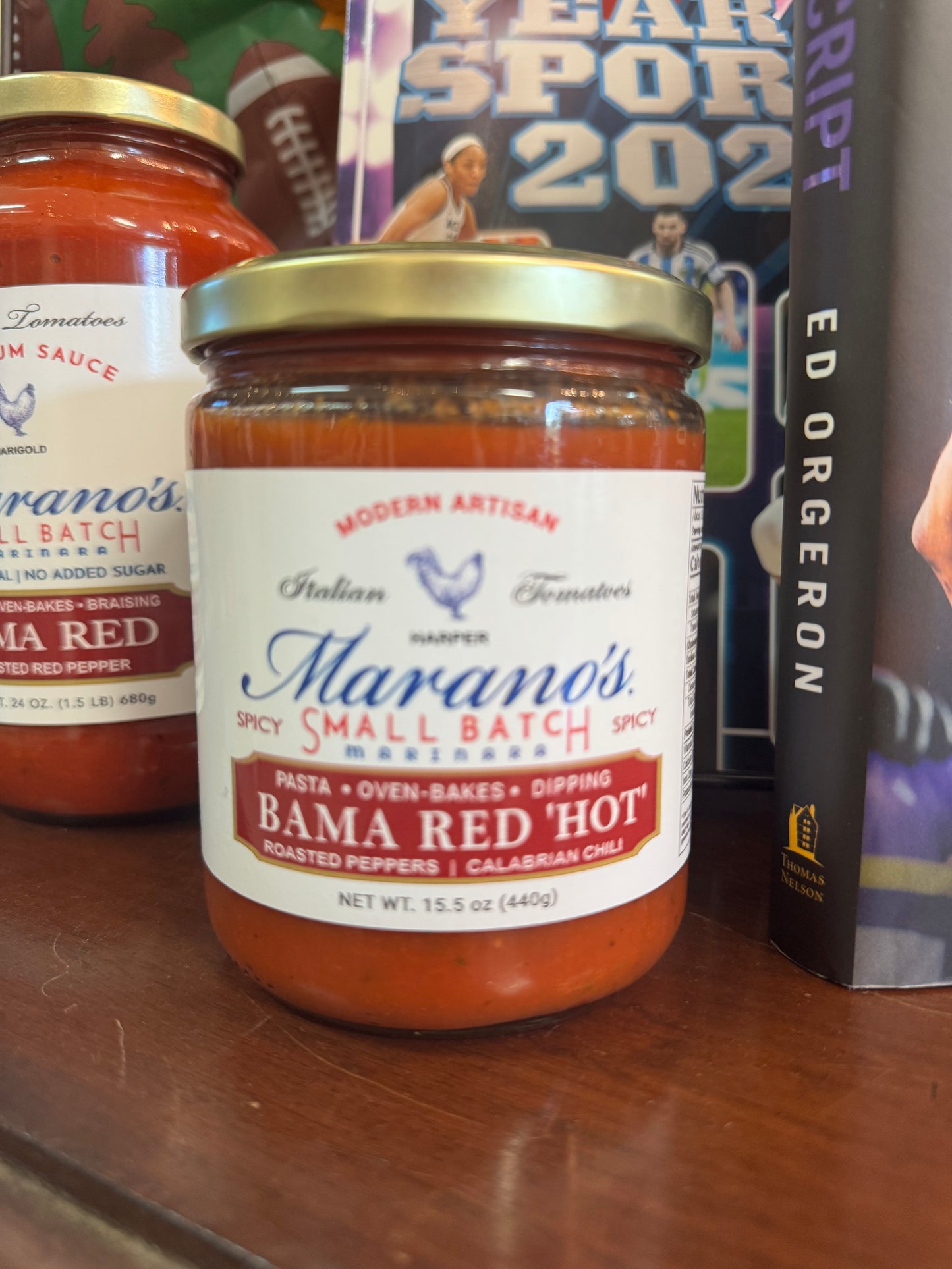 Jar of Marano's Spicy Small Batch Bama Red Hot sauce on a shelf with sports-themed books.