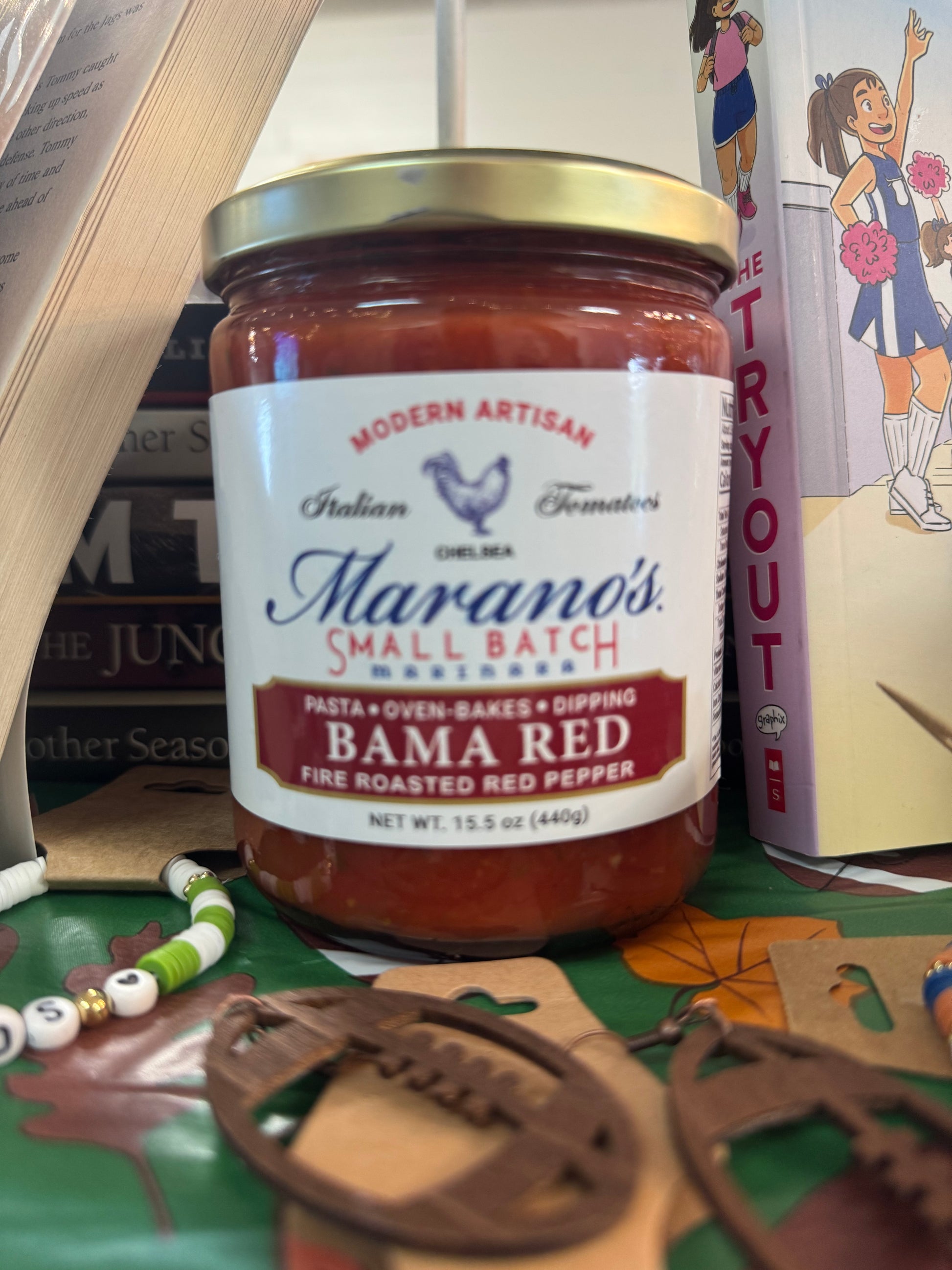 Jar of Marano's Spicy Small Batch Bama Red sauce on a shelf with sports-themed books.