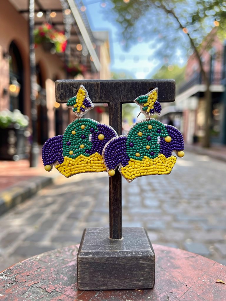 Beaded Mardi Gras jester hat earrings in purple, green, and gold displayed on a wooden stand with a festive street background