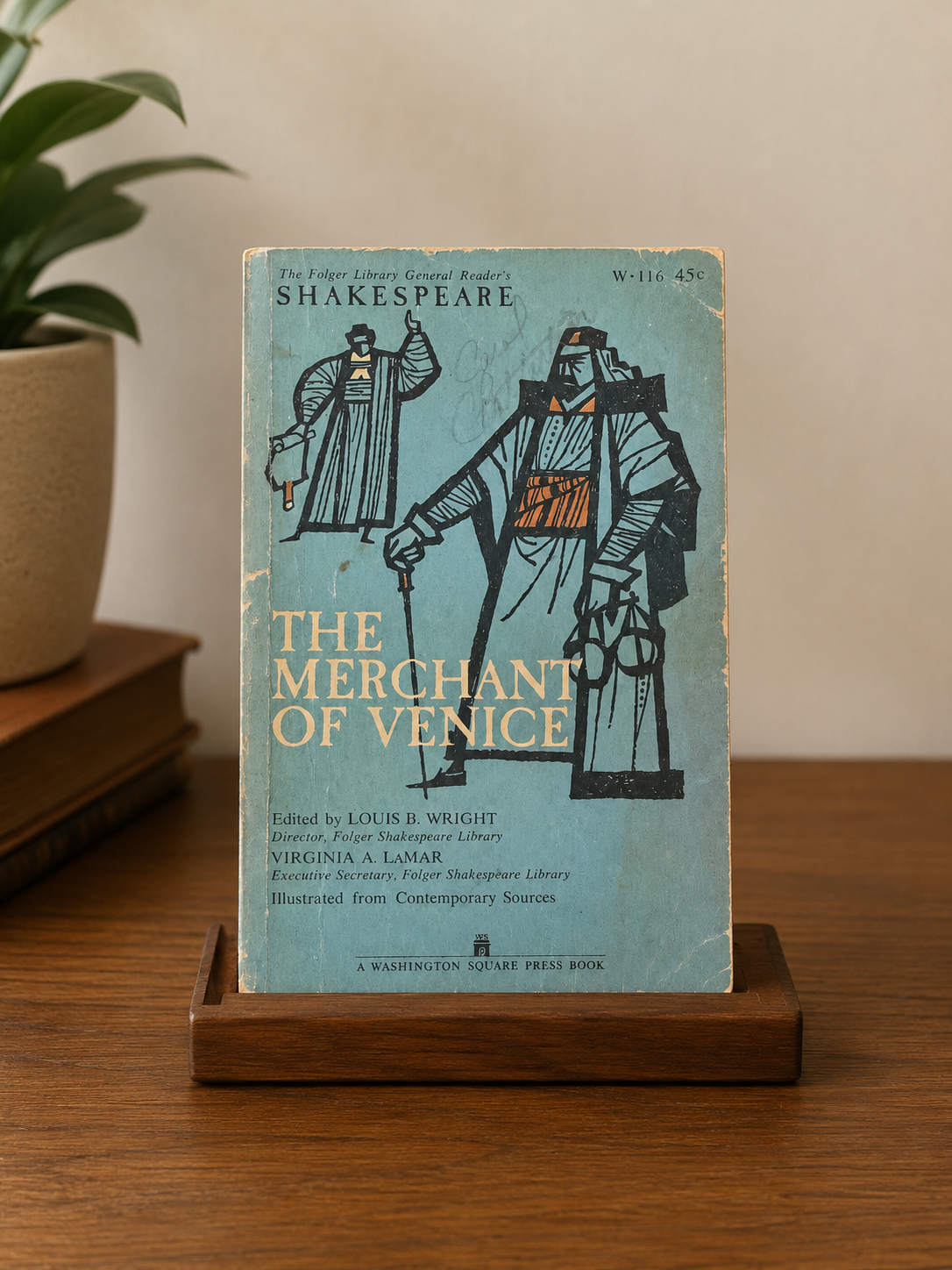 Book titled 'The Merchant of Venice' by William Shakespeare on a wooden stand with a plant in the background.