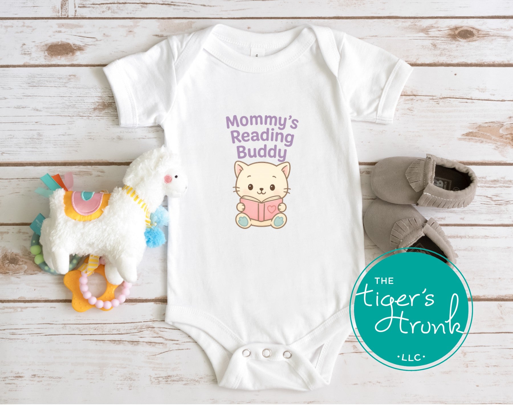 White baby bodysuit with cute pastel kitten holding a book and text Mommy’s Reading Buddy, styled with baby shoes and toy.