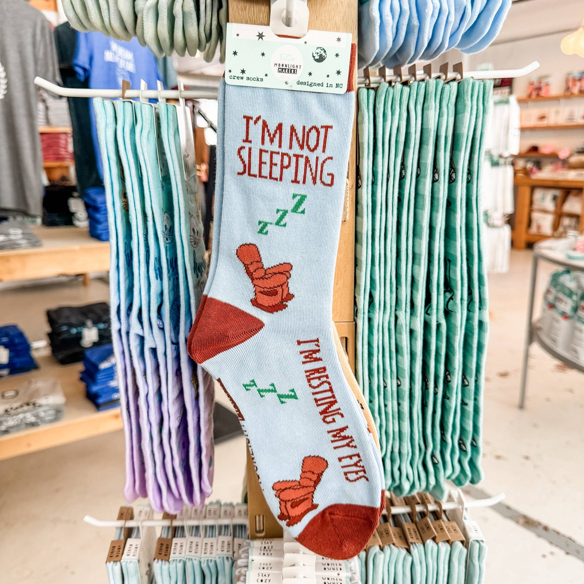 Light blue novelty crew socks that read I’m Not Sleeping, I’m Resting My Eyes hanging on a retail display rack inside a gift shop, with stacks of other socks in the background.