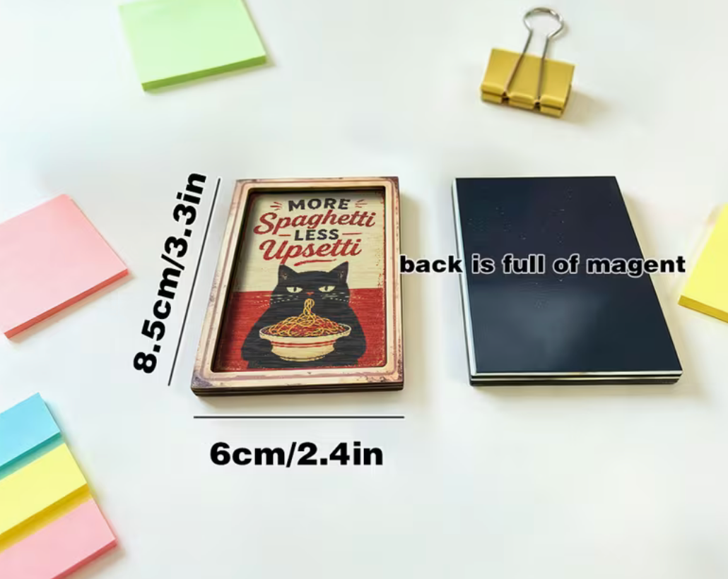 A rectangular printed magnet or plaque featuring a black cat eating spaghetti with the text 'MORE Spaghetti LESS Upset!' is shown on a white surface with dimension labels. A stack of dark magnets sits beside it, and sticky notes and binder clips are arranged around it as props.