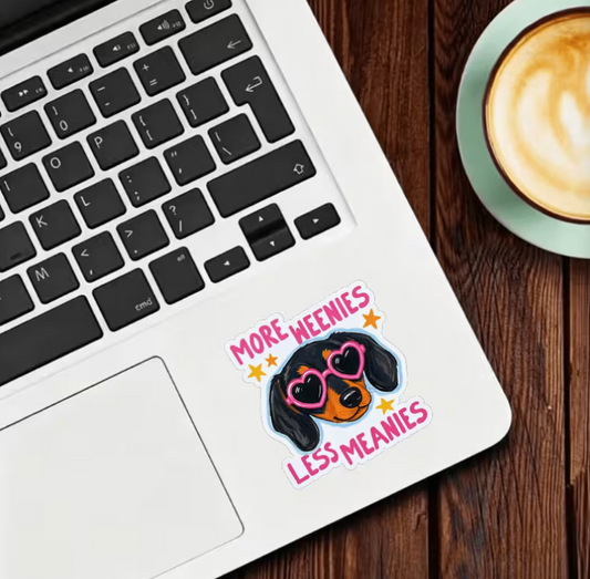 More Weenies Less Meanies dachshund sticker featuring a black and tan wiener dog wearing pink heart-shaped sunglasses with colorful stars and pink lettering on a laptop