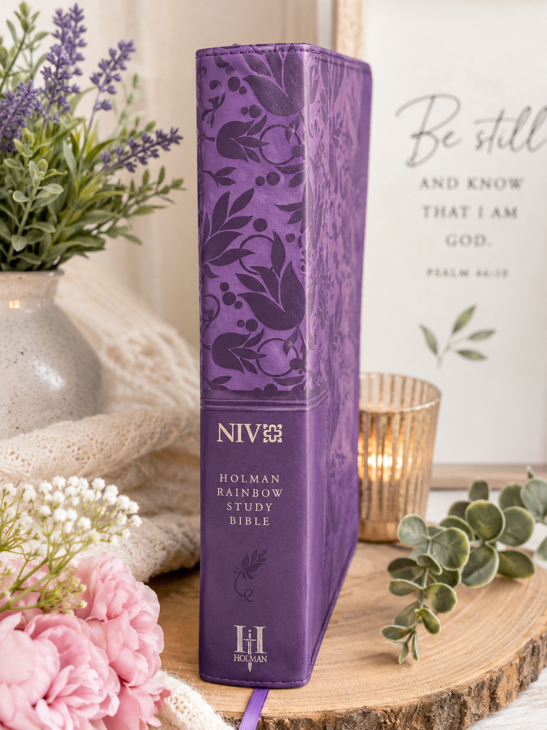 Purple NIV Holman Rainbow Study Bible displayed upright with the spine facing forward on a rustic wood surface, styled with lavender flowers, a candle, greenery, and a framed Bible verse in the background.