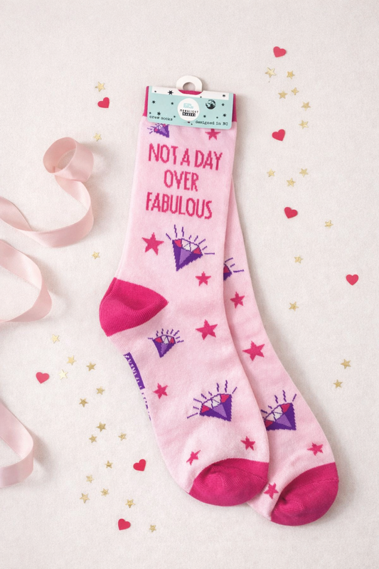 Flatlay of pink Not a Day Over Fabulous novelty crew socks in retail packaging with purple diamond and red star design, styled with pink ribbon, heart confetti, and gold stars on a light background for gift display and product listing
