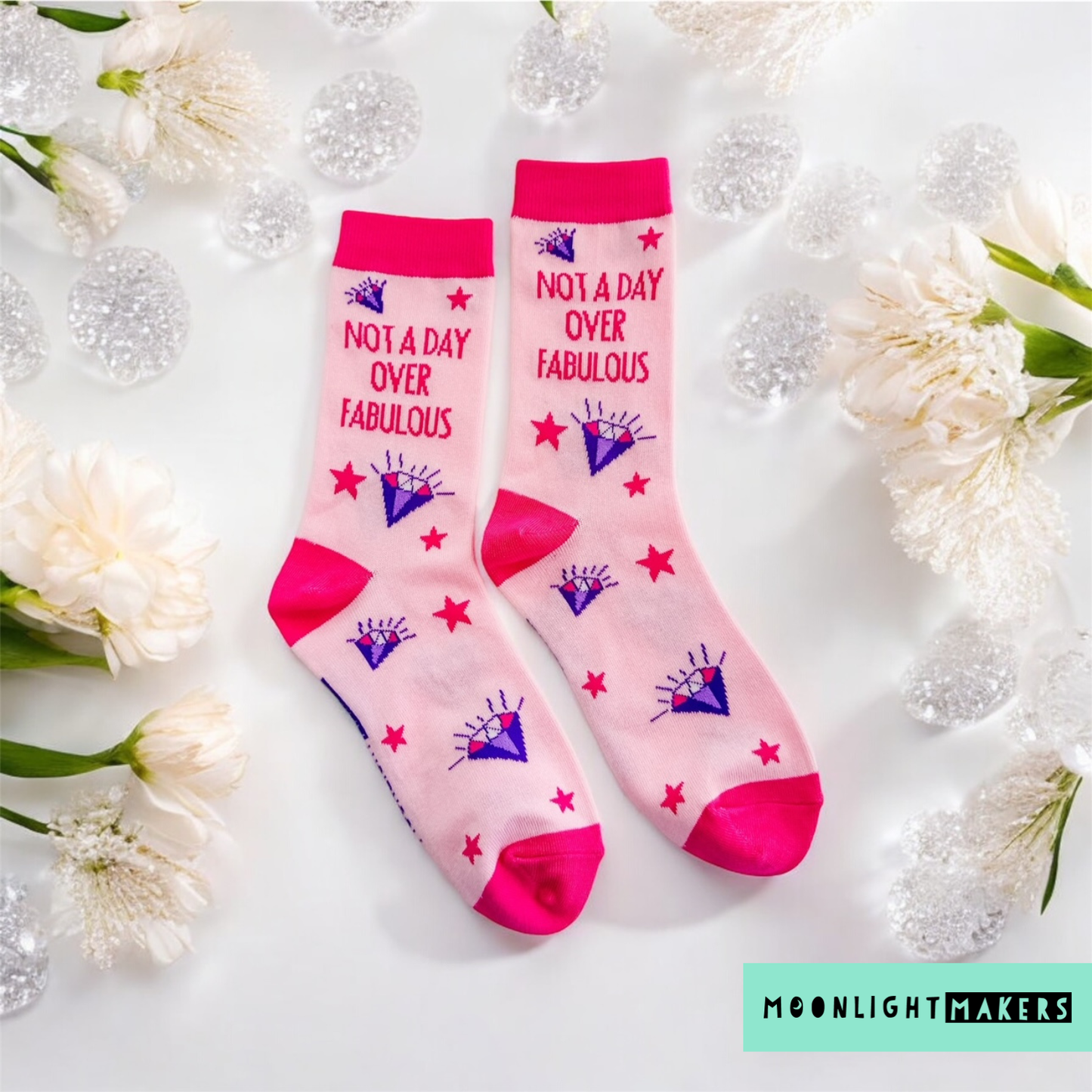 Flatlay of pink Not a Day Over Fabulous novelty crew socks with purple diamond and red star design, styled on a white background with white flowers and clear decorative accents, brand watermark visible in the corner for product listing display