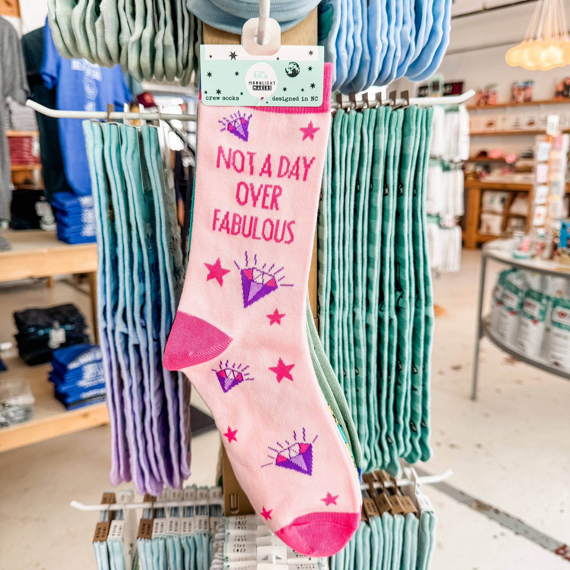 Pink Not a Day Over Fabulous novelty crew socks with purple diamond and red star design hanging on a retail display rack, shown in store with Moonlight Makers packaging visible and surrounding socks in soft pastel colors.