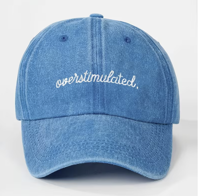 A blue denim-textured baseball cap with the word 'overstimulated.' embroidered in white script across the front, shown against a plain light background.