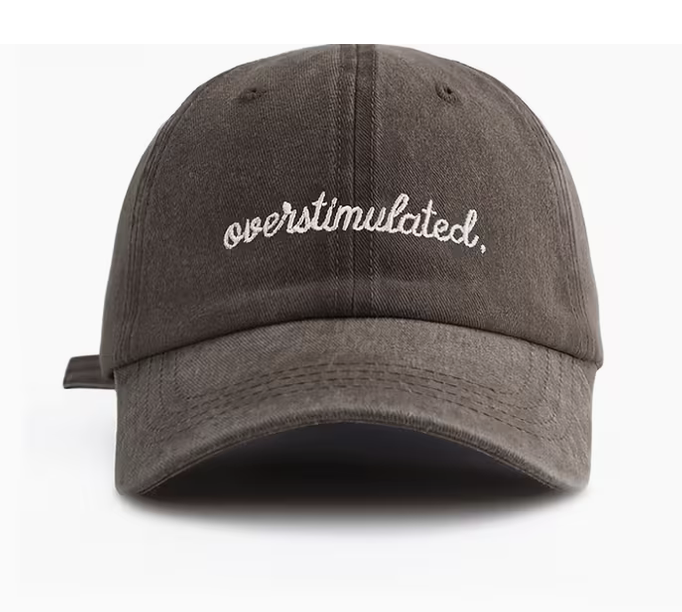 A dark brown washed cotton baseball cap with 'overstimulated.' embroidered in white script on the front, shown against a plain white background.
