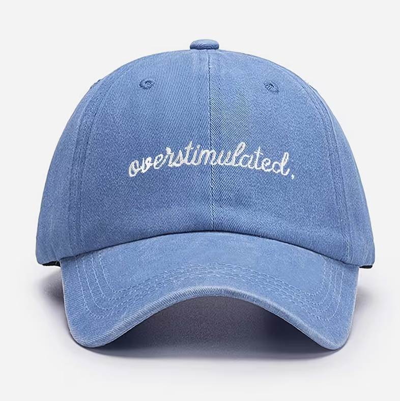 A light blue washed cotton baseball cap with 'overstimulated.' embroidered in white script across the front, shown against a plain white background.