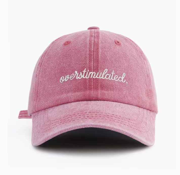 A pink washed cotton baseball cap with 'overstimulated.' embroidered in white script across the front, shown against a plain white background.