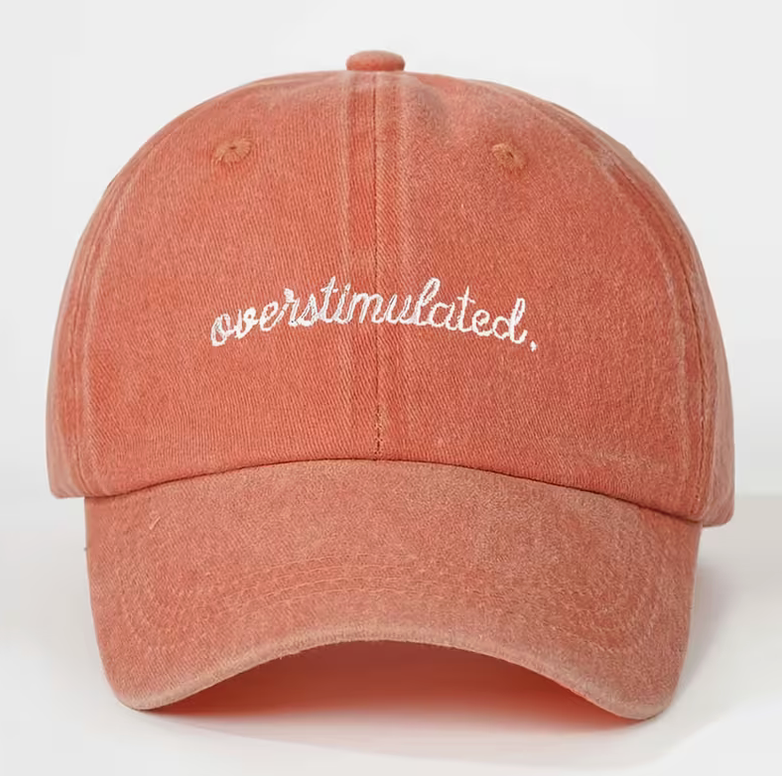A washed terracotta pink baseball cap with 'overstimulated.' embroidered in white script across the front, shown against a plain white background.