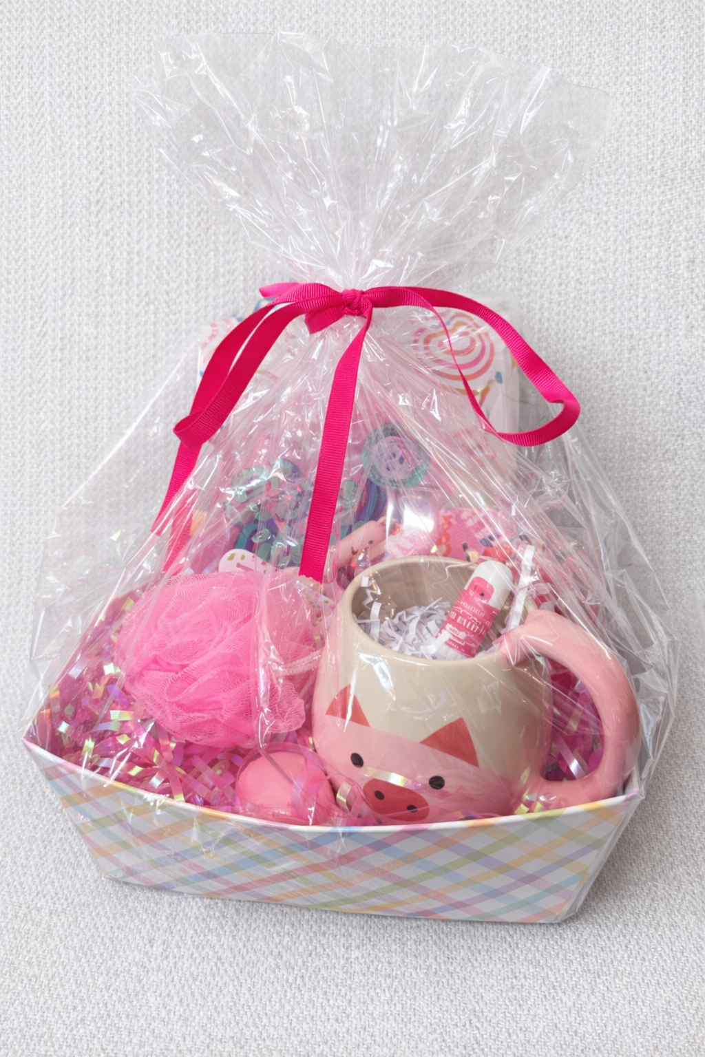 Pink gift basket with pig mug, bath sponge, beauty items, and small treats wrapped in clear gift wrap with bright pink ribbon