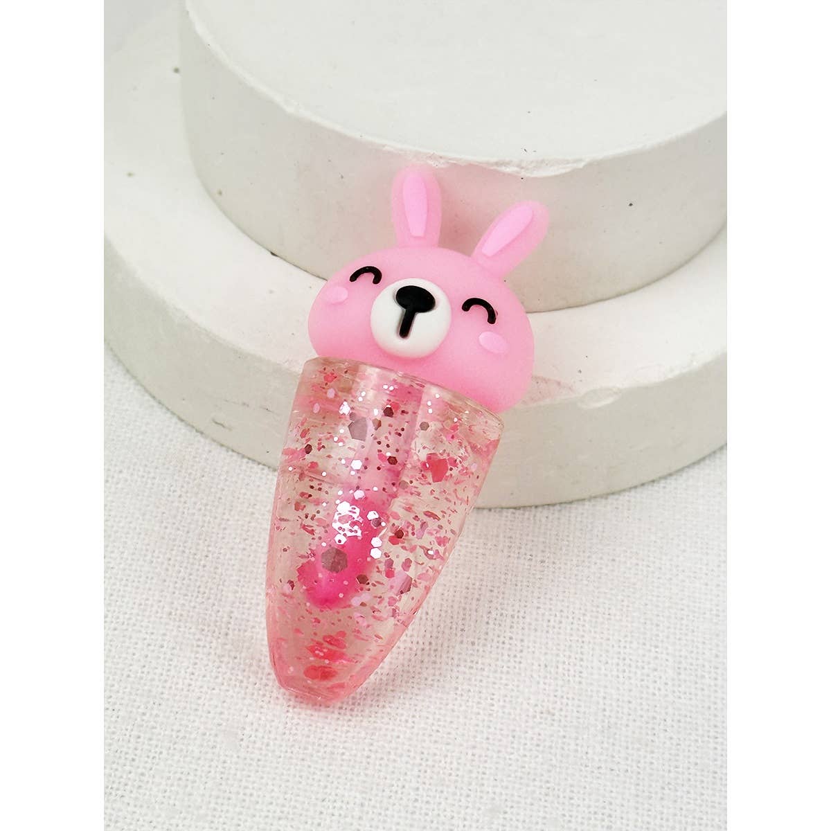 Single pink rabbit lip oil with glitter-infused clear oil in a carrot-shaped tube, featuring a smiling bunny cap, displayed on a neutral background