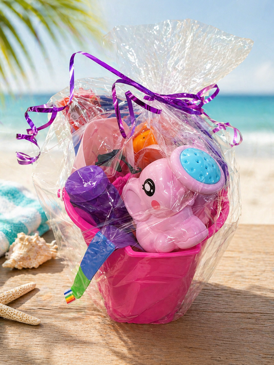 Pink summer kids gift basket in a sand pail filled with bubbles, sand toys, water toys, pool float, comet ball, and outdoor play items wrapped in clear gift wrap with ribbon on a beach background