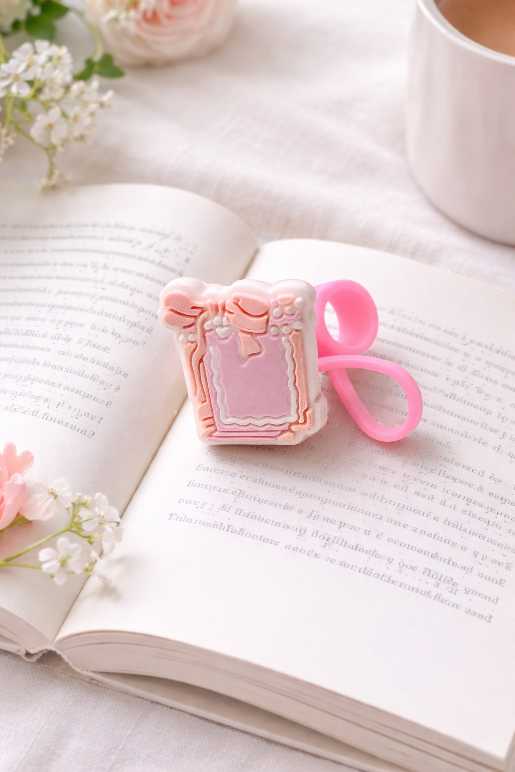 Pink vintage-style book silicone straw topper with bow detail resting on an open book beside flowers and a white mug.