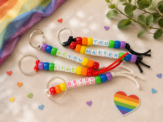 Rainbow beaded pride keychains with positive messages displayed on a soft neutral flat lay background with subtle rainbow accents, greenery, and colorful heart confetti