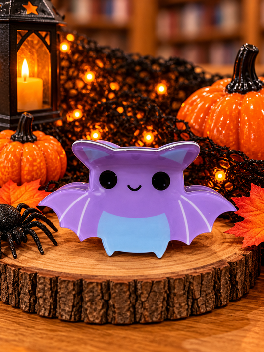 Cute purple bat Halloween hair clip measuring approximately 3 inches, featuring outstretched wings, black eyes, a smiling face, and a light blue belly, displayed with pumpkins and festive Halloween decorations.