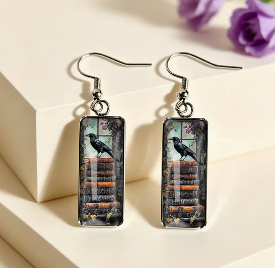 Rectangular dangle earrings featuring a black raven perched on a stack of antique books with dark gothic artwork