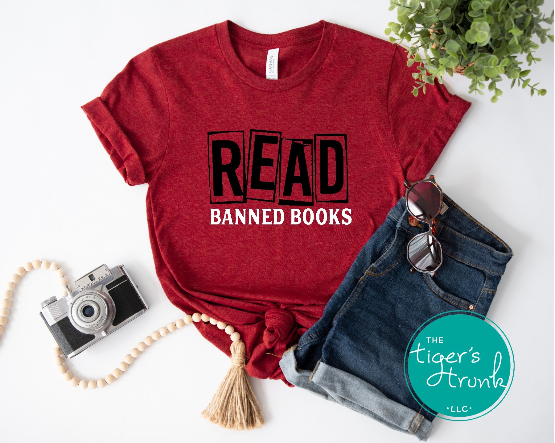 Red t-shirt with 'READ BANNED BOOKS' text, sunglasses, camera, and denim shorts on a white background.