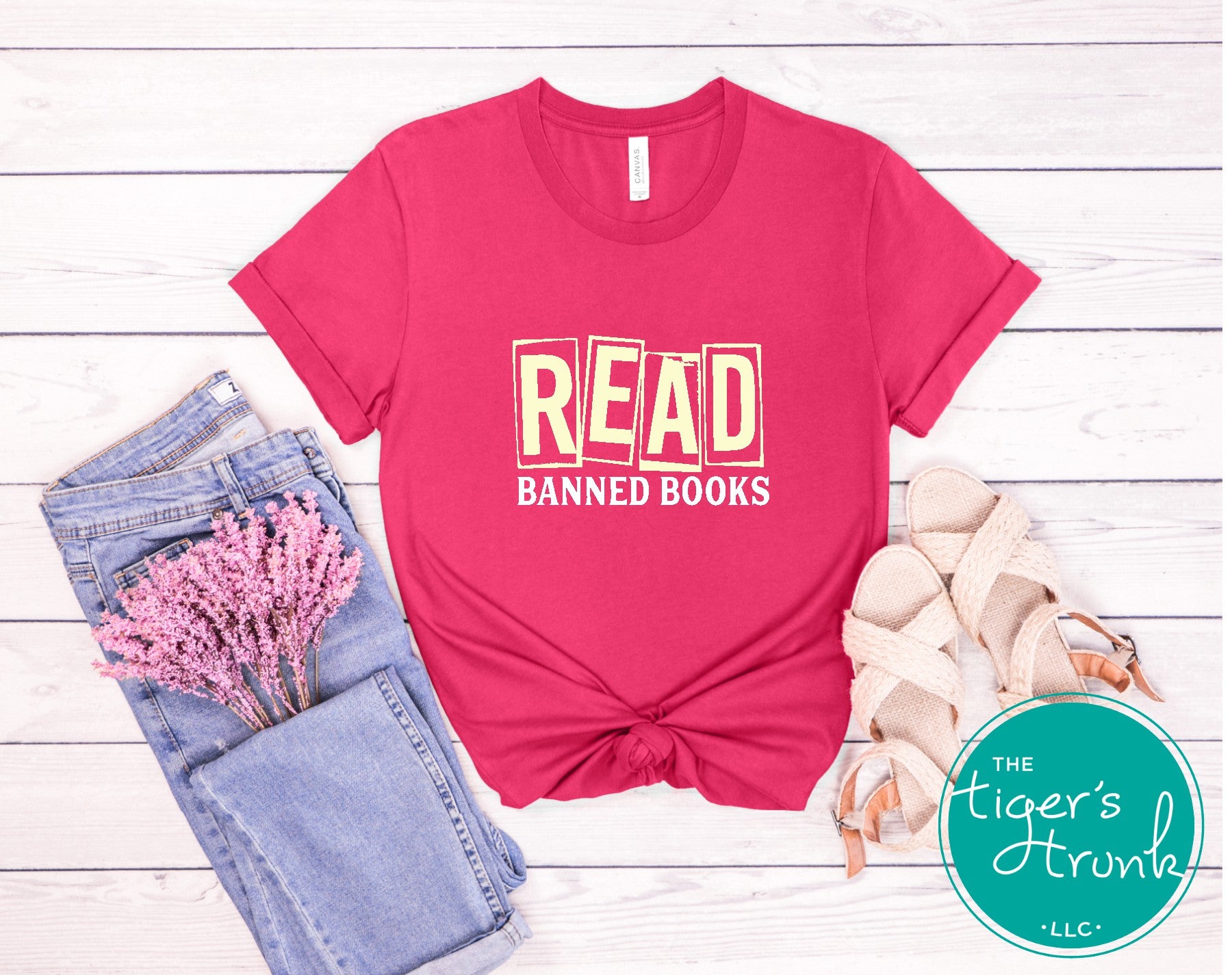 Pink t-shirt with 'READ BANNED BOOKS' text on a wooden surface with jeans and flowers.